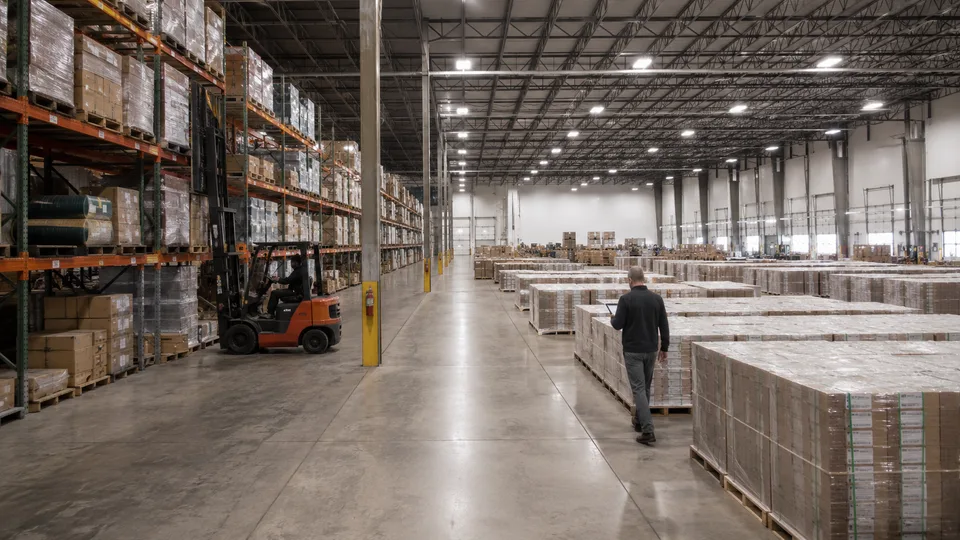 Warehouse operator with a tablet walking past shrink-wrapped project pallets beside loaded racking