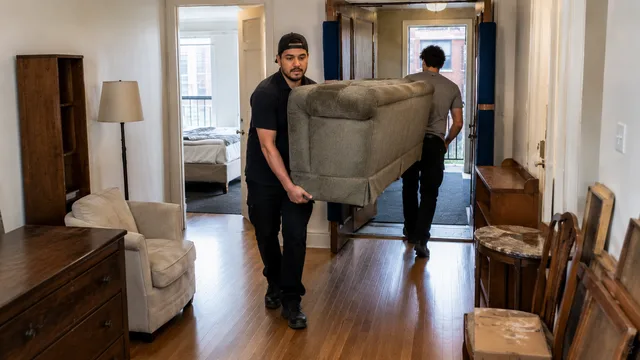 Movers carrying a used sofa out of a furnished apartment past a dresser and side tables