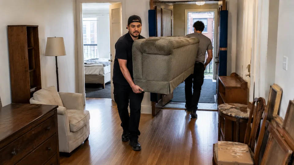 Movers carrying a used sofa out of a furnished apartment past a dresser and side tables